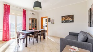 A modern living room with a dining area, a grey sofa, a table with magazines, and a painting on the wall.