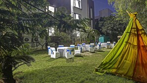 A grassy outdoor area with white chairs with blue sashes, a green tent, and buildings in the background. - Dormy Villa By Dreamz Hospitality (Gurugram)