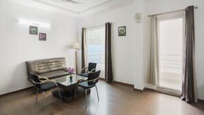 A spacious living room with a sofa, two chairs, a coffee table, a ceiling fan, and large windows with curtains. - Dormy Villa By Dreamz Hospitality (Gurugram)