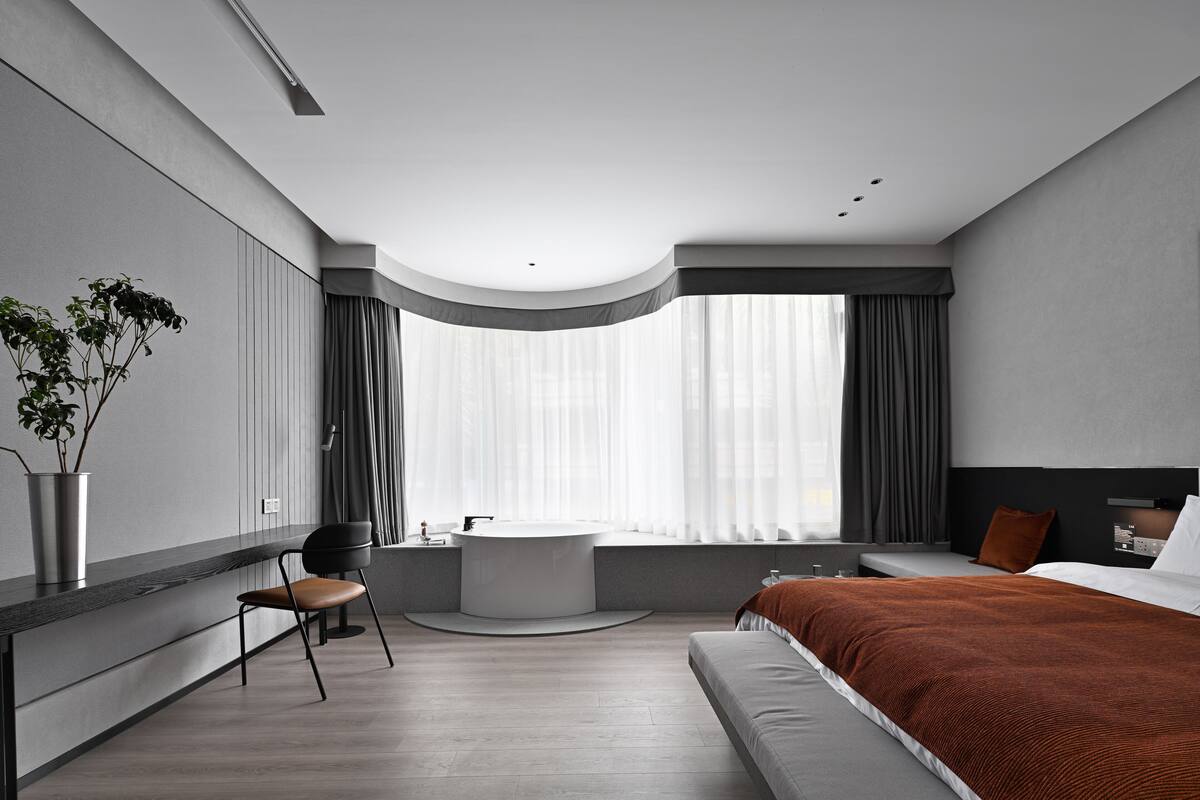 A modern hotel room with a large bed, a round bathtub, a seating area with a chair, a small table, and a large window with curtains.