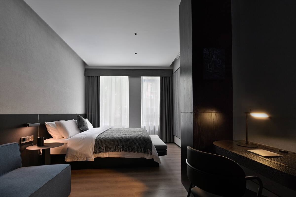 A modern hotel room with a large bed, bedside tables, a desk with a lamp, and a view of the city through large windows.
