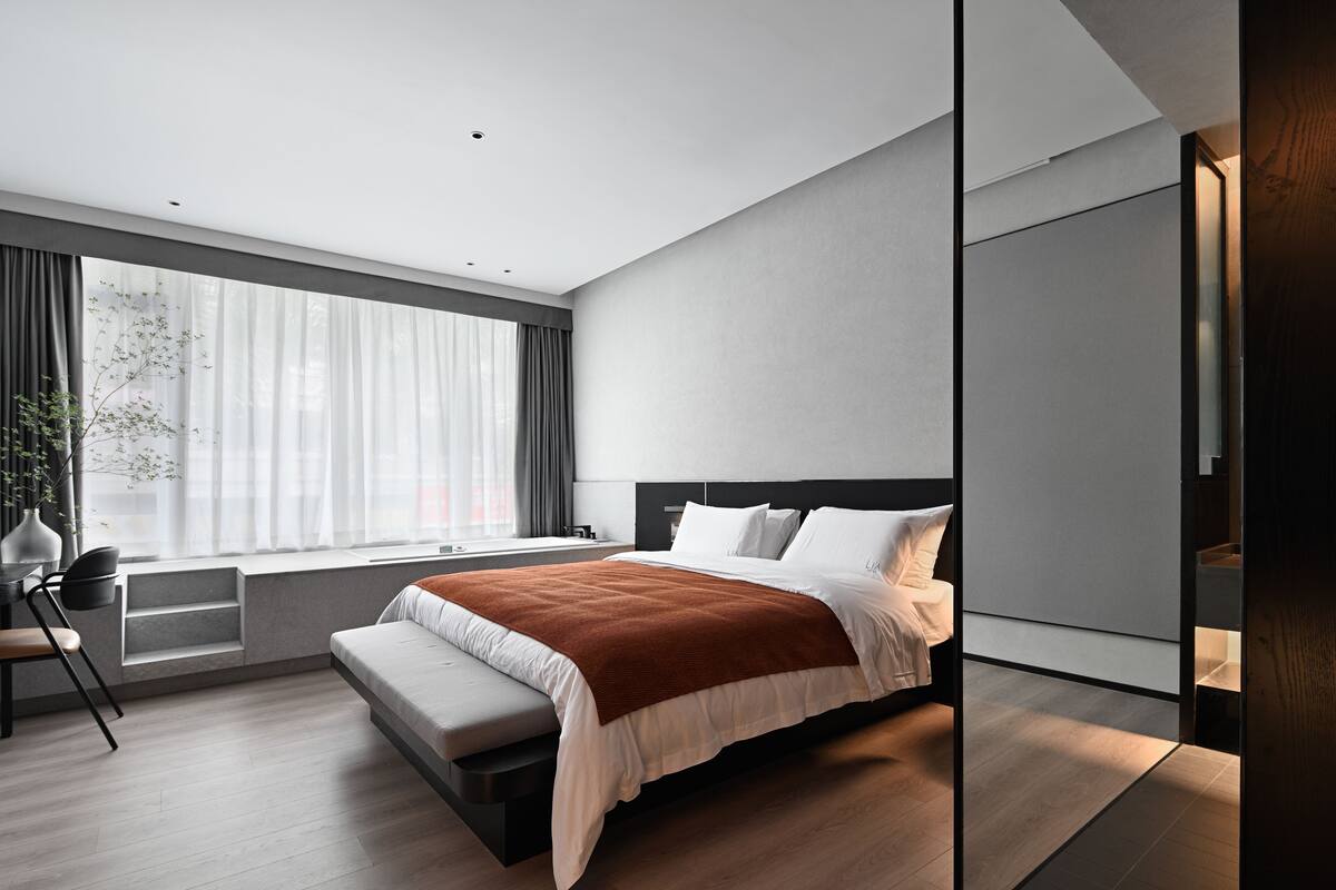 A modern bedroom with a large bed, a bench, a desk, and a window with curtains.