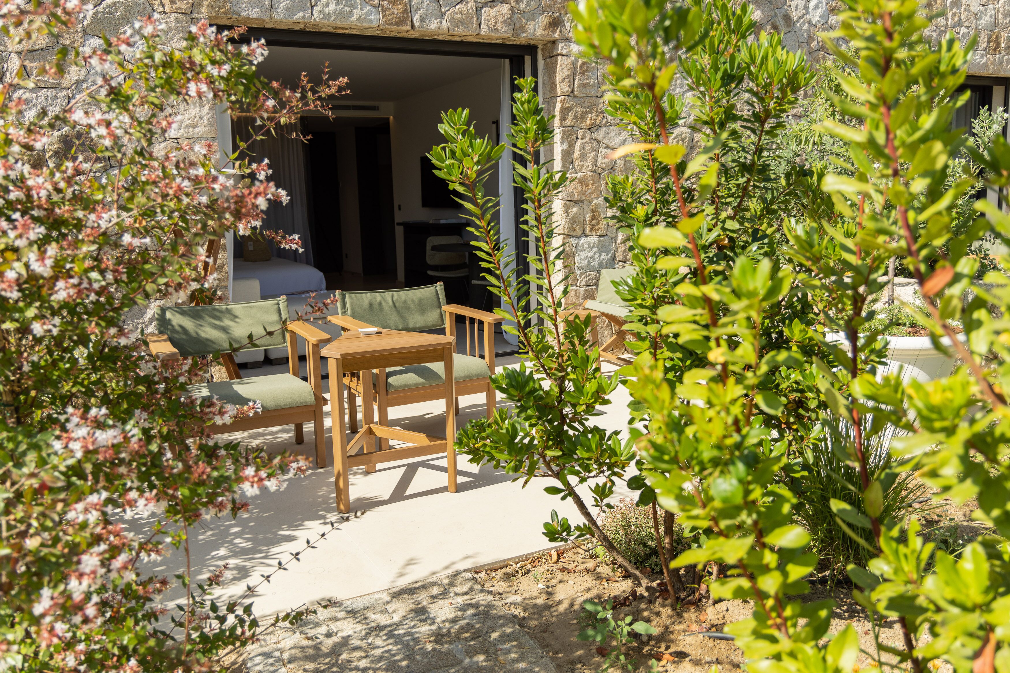 An outdoor seating area with a table and chairs, surrounded by greenery and a stone wall.