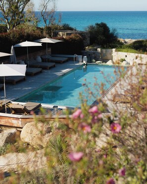 Zona de piscina con sillones de descanso y sombrillas, una pared de piedra y vistas al mar.