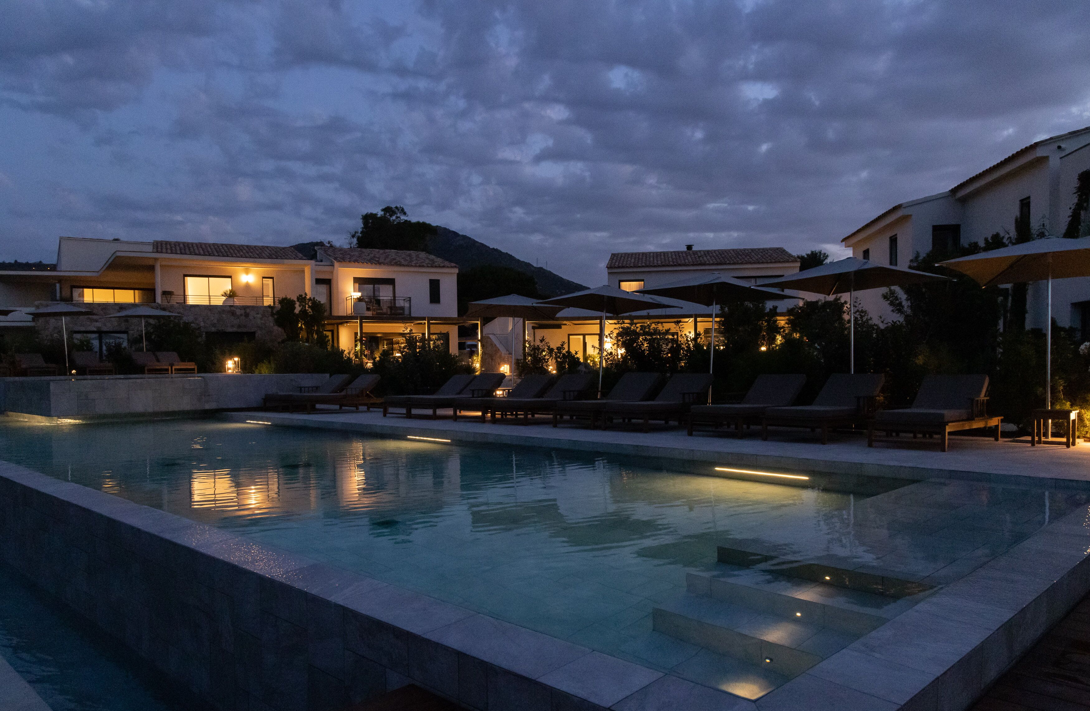 A swimming pool with lounge chairs and umbrellas, surrounded by buildings with lights on.