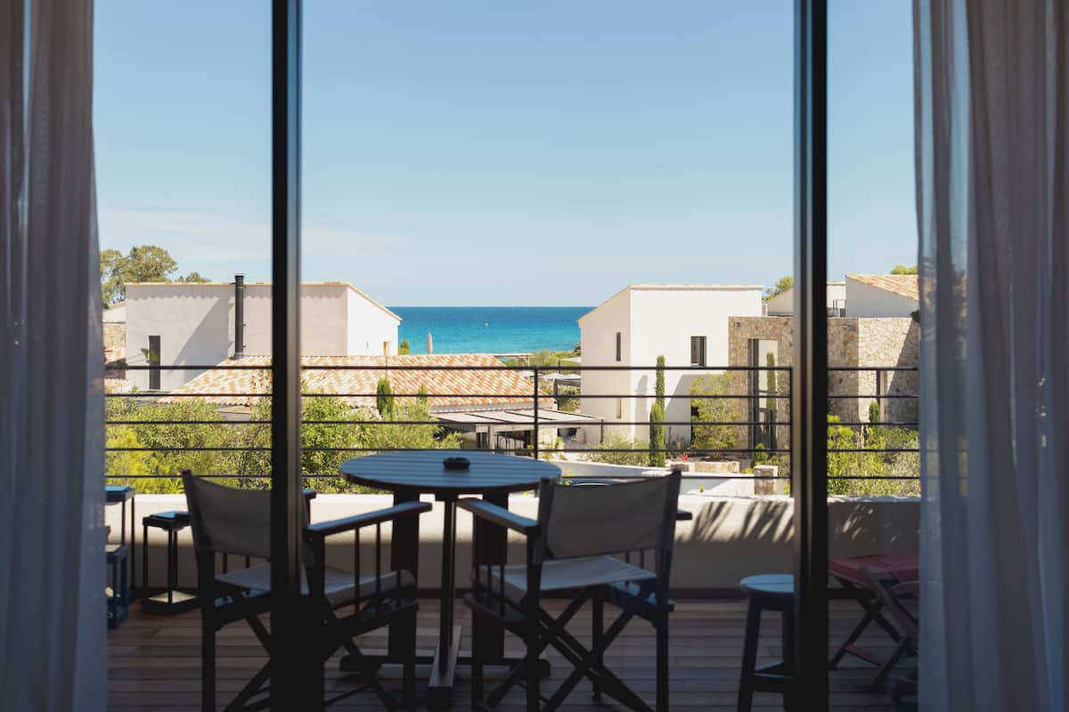An outdoor patio with a round table and chairs, overlooking a coastal area with buildings and the sea.
