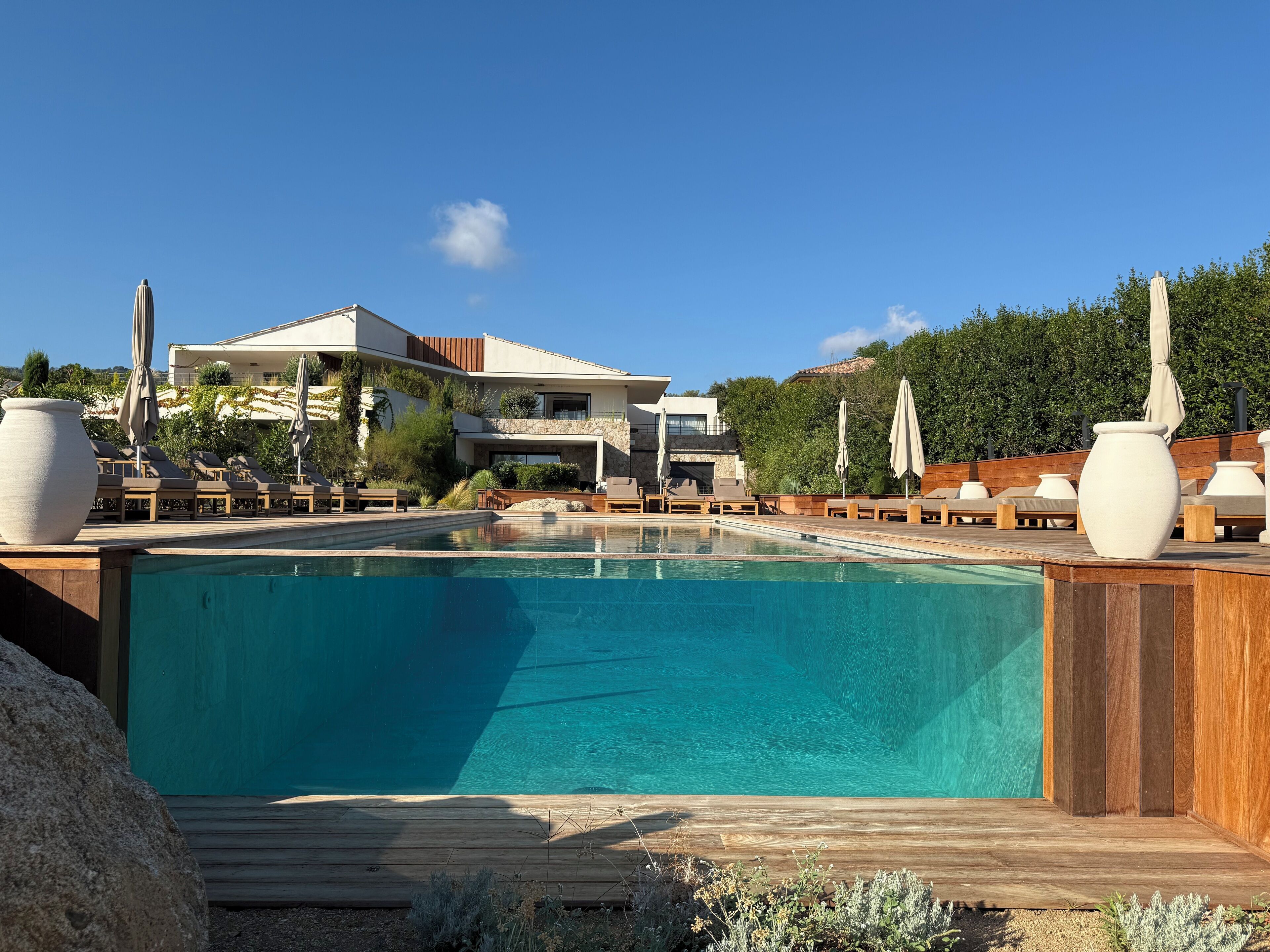 A modern pool area with lounge chairs and umbrellas, surrounded by a wooden deck and a stone building in the background.