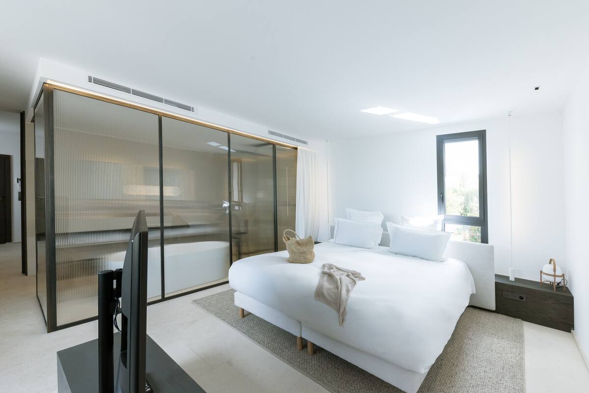 A modern bedroom with a large bed, a TV, and a sliding glass door leading to a bathroom.