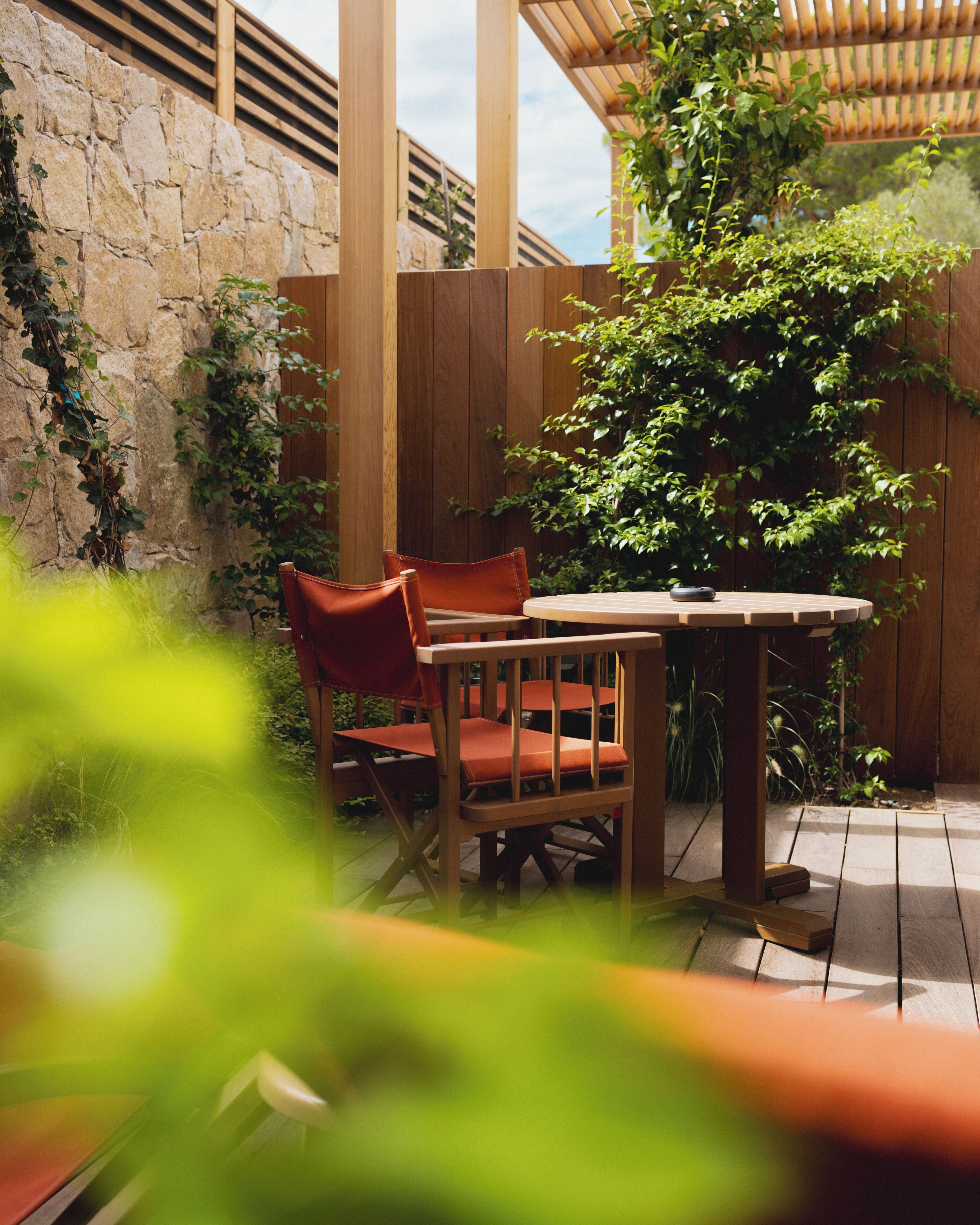 A wooden table and two chairs in an outdoor patio area with a stone wall and wooden fence.