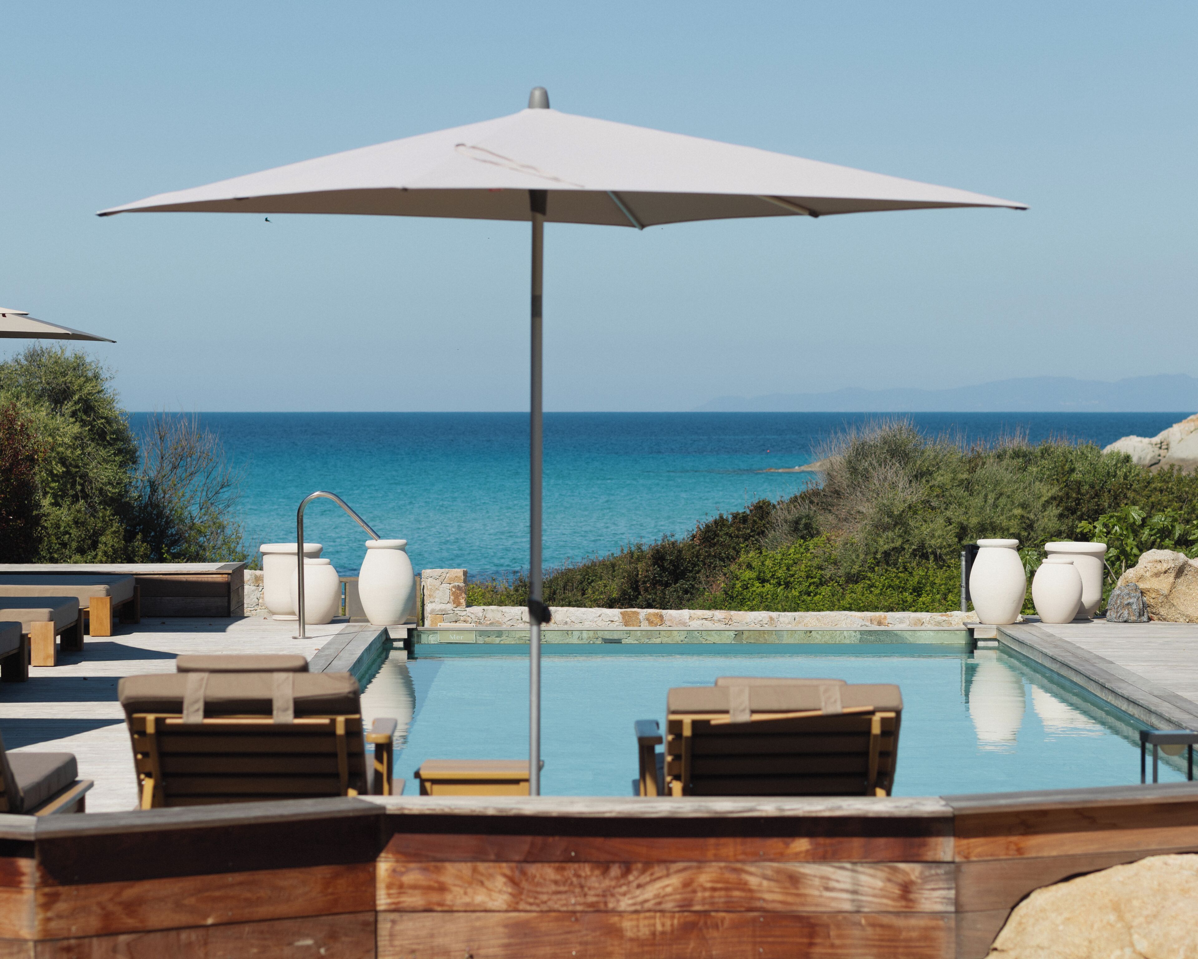 A pool area with lounge chairs, a large umbrella, and a view of the sea.