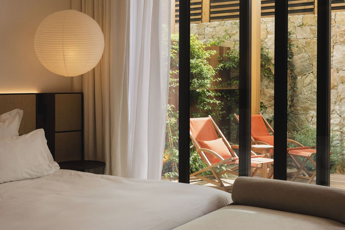 A bedroom with a bed, a hanging lamp, and a view of an outdoor area with chairs and a wooden structure.
