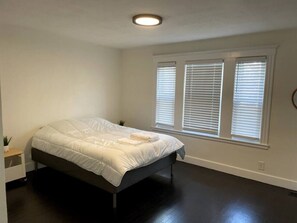 Une chambre à coucher avec un lit, une table de chevet, une fenêtre avec des stores et un luminaire au plafond.