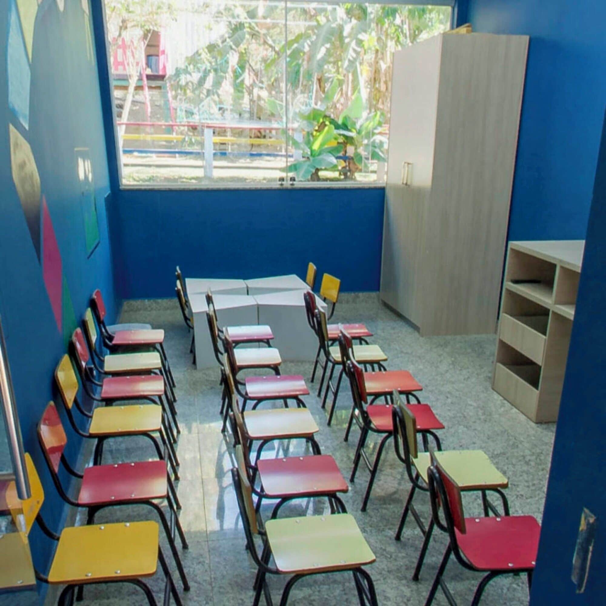 Un aula con pupitres y sillas de colores dispuestos en filas, un ventanal con vista a la vegetación y una pared azul.