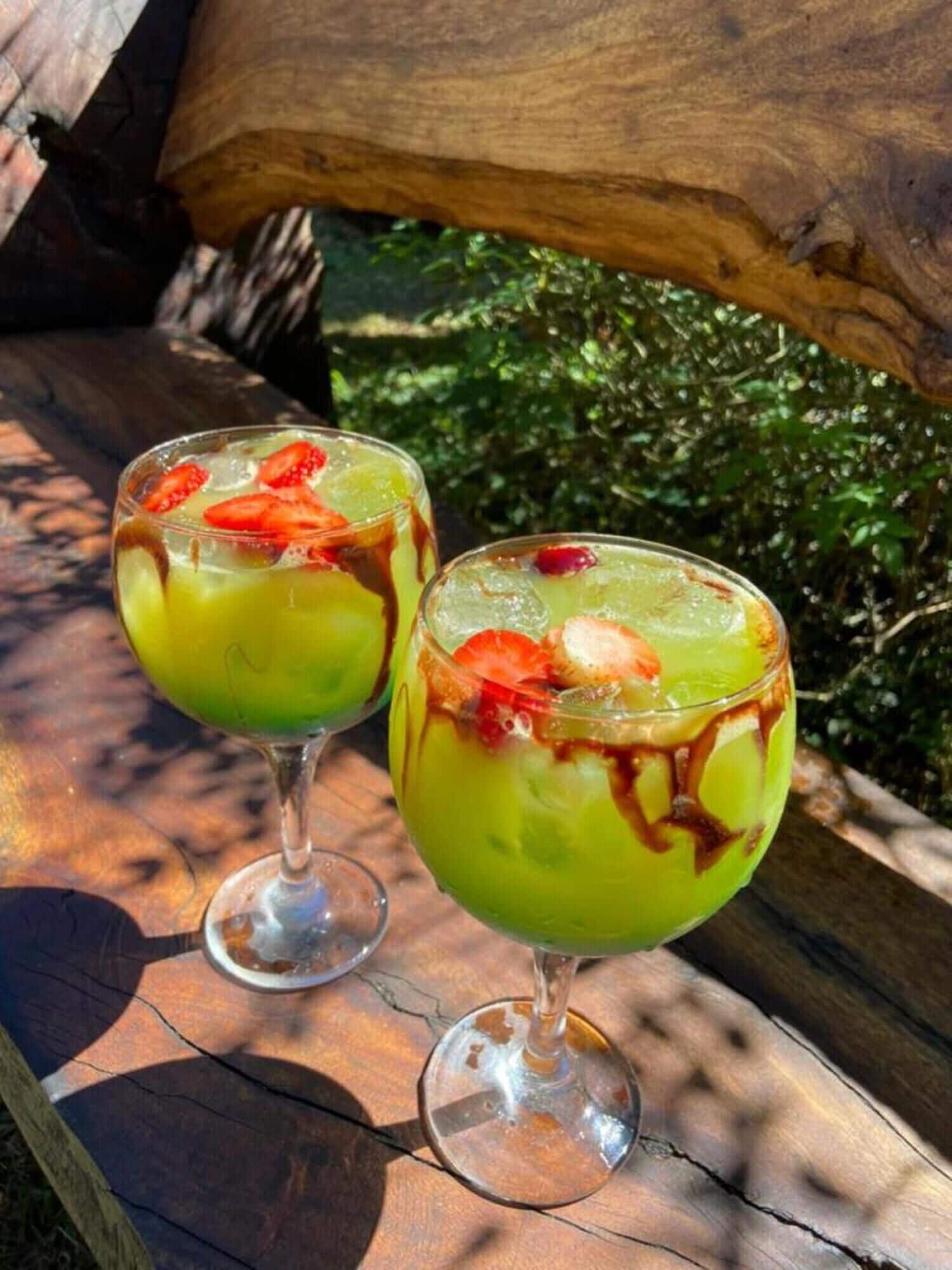 To glas med en grøn drik, jordbær og en brun sirup, placeret på en træoverflade udendørs.
