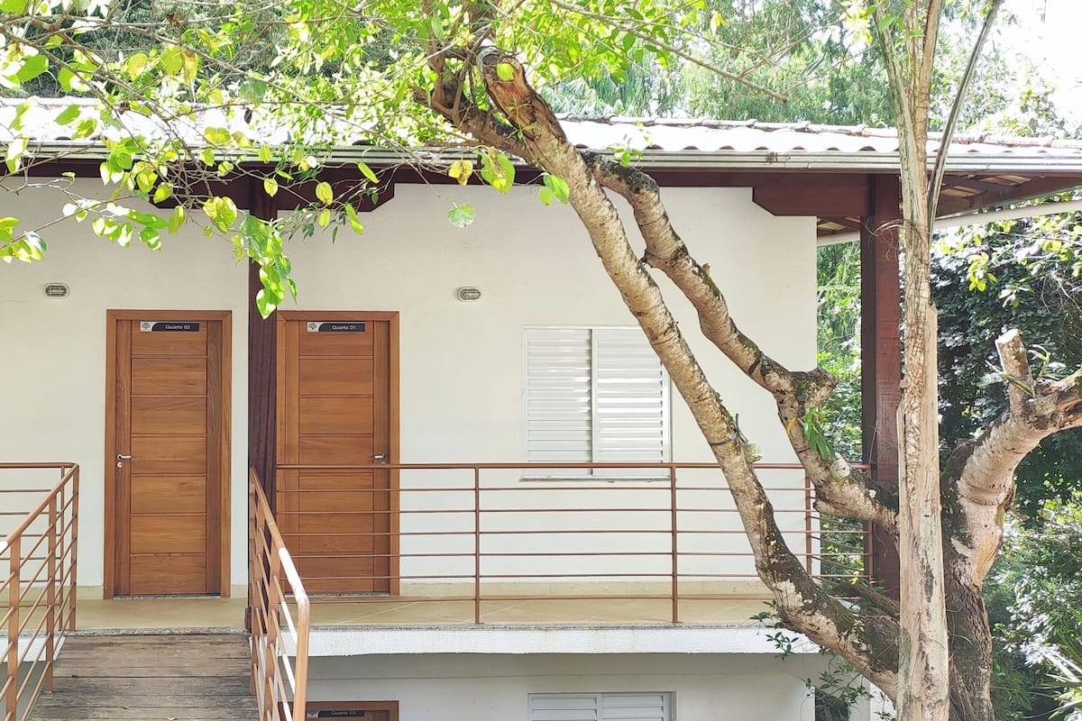 Un bâtiment à deux étages, doté d’un escalier en bois menant à un balcon. On y trouve deux portes en bois à l’étage supérieur, et le bâtiment est entouré d’arbres.