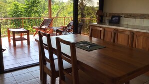 Une table de salle à manger en bois avec des chaises, un coin cuisine avec un mur recouvert de carreaux, et un balcon offrant une vue sur une végétation luxuriante.