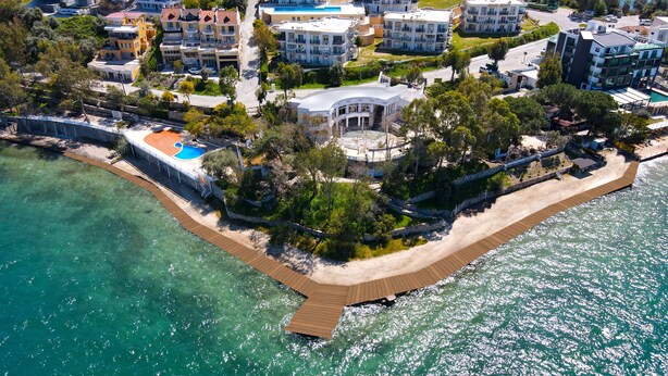An aerial view of a coastal property with a wooden deck, clear turquoise waters, and modern buildings with balconies. - Roseira Beach Resort (Milas)