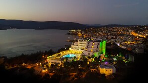 A coastal cityscape at dusk with illuminated buildings, a marina, and a large hotel with a pool and outdoor area. - Roseira Beach Resort (Milas)