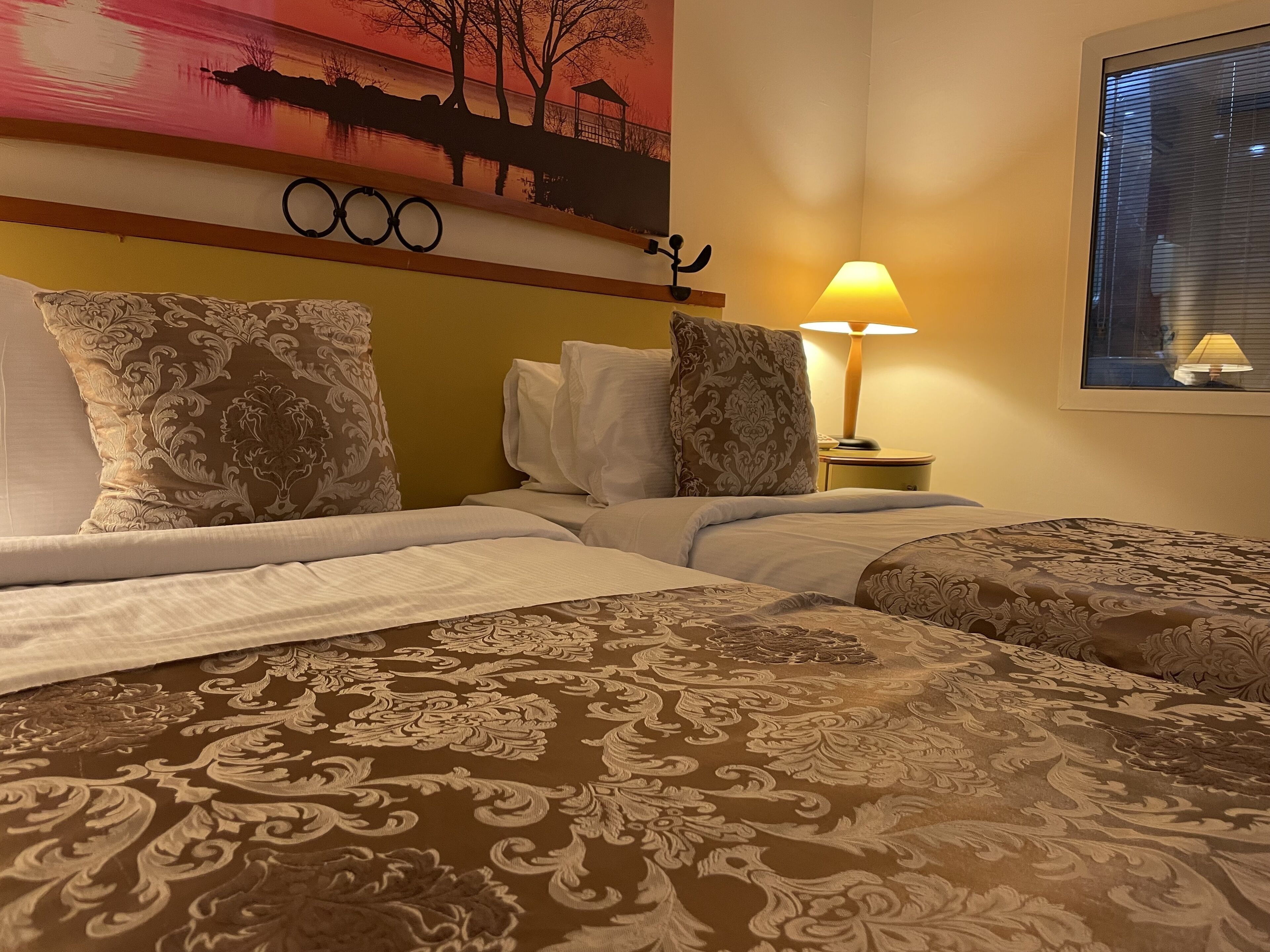 Habitación de hotel con dos camas, un cuadro de un atardecer, una lámpara y una ventana con persianas.
