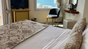 A hotel room with a bed, a television, a chair, a desk, a window with a view of the sea, and a mirror with lights. - Roseira Beach Resort (Milas)