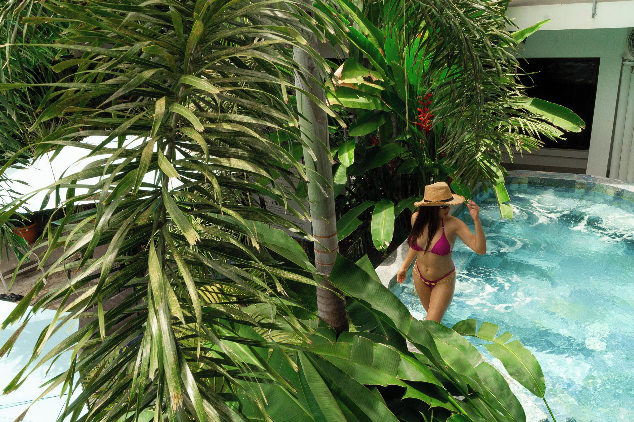 Mulher de biquíni e chapéu perto de uma piscina, rodeada por plantas tropicais.