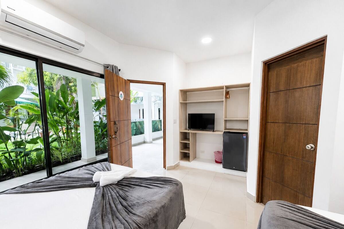 A bedroom with two beds, a TV, a refrigerator, and a large window with plants.