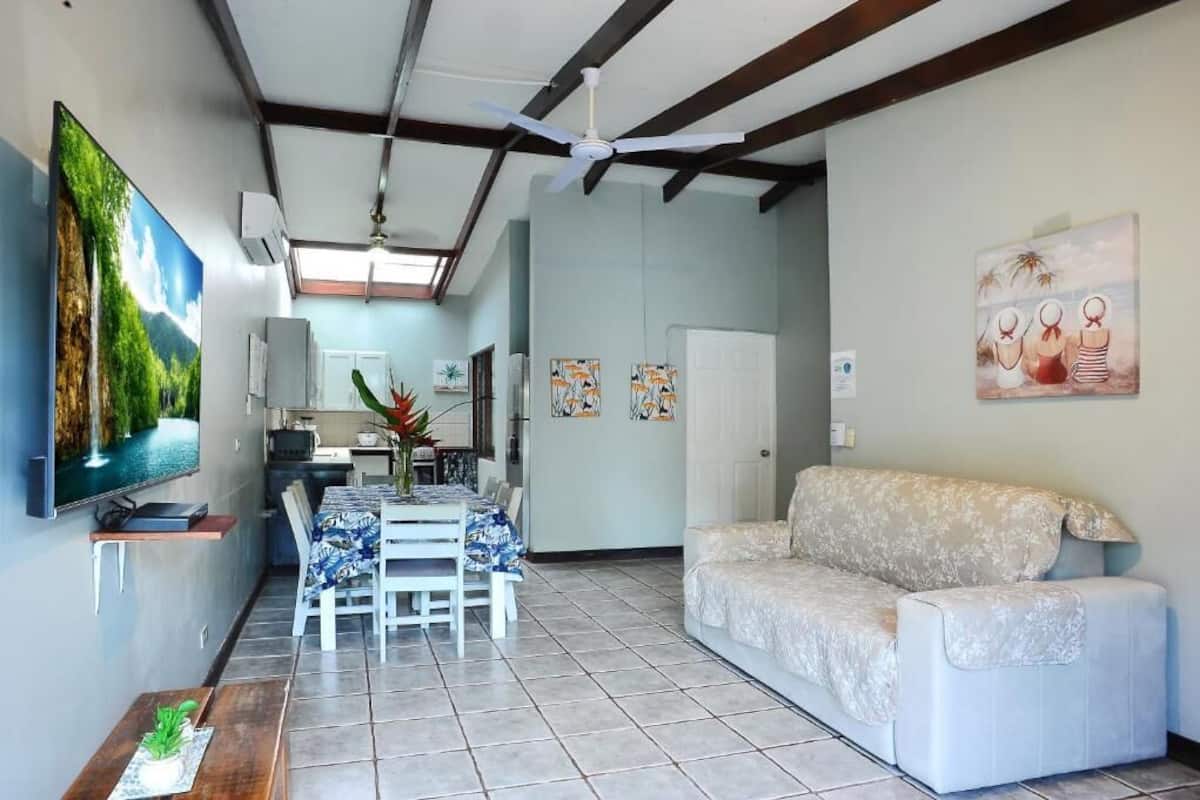 A living room with a TV, sofa, dining table, and kitchen in the background.