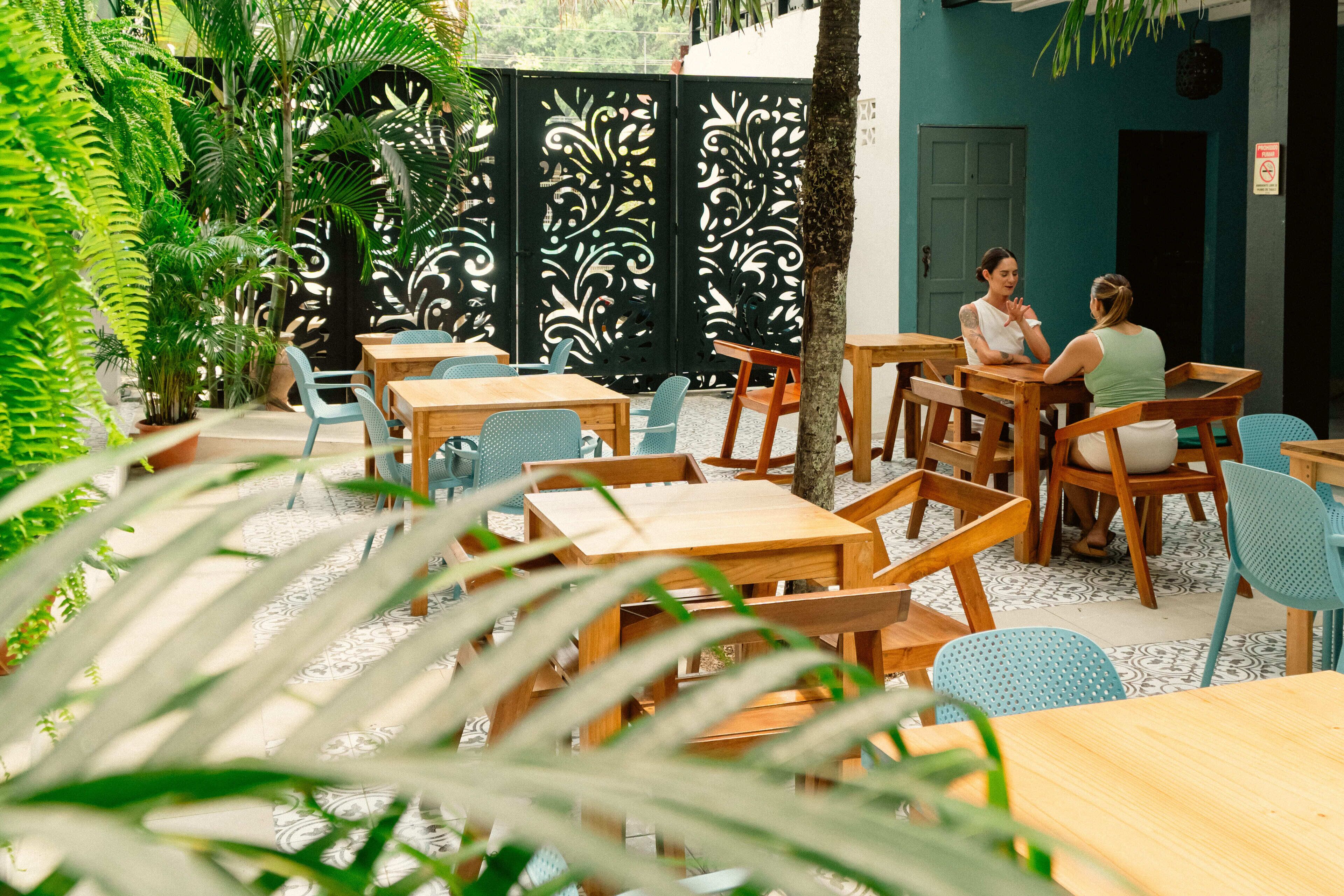 An outdoor seating area with wooden tables and chairs, surrounded by potted plants and a decorative black fence.