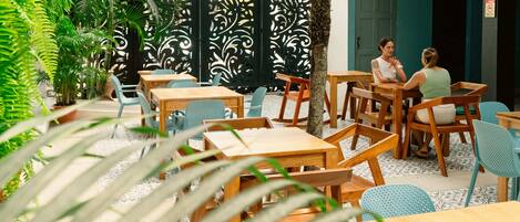 An outdoor seating area with wooden tables and chairs, surrounded by potted plants and a decorative black fence.