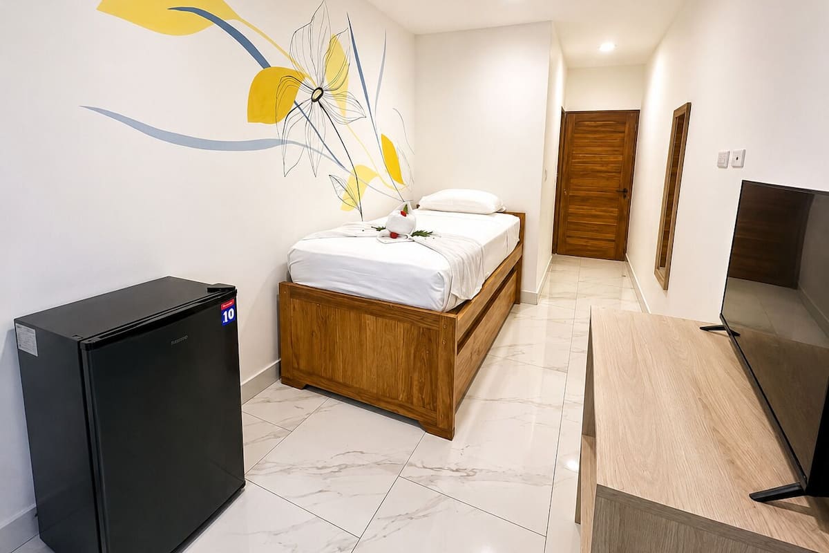 A bedroom with a bed, a small black refrigerator, a wooden desk, a TV, and a wall with abstract art.