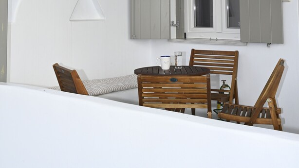 An outdoor seating area with a table and chairs, a window with shutters, and a view of a hillside. - Τinos Deluxe Home (Τήνος)