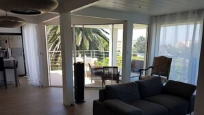 A living room with a sofa, a dining table with chairs, and a balcony with a view of palm trees. - Very pretty 4* listed wooden house in Biarritz with swimming pool (Biarritz)