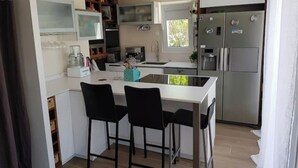 A modern kitchen with a central island, two black bar stools, a built-in oven, a refrigerator, and a window providing natural light. - Very pretty 4* listed wooden house in Biarritz with swimming pool (Biarritz)