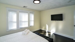 A bedroom with a large bed, two windows with blinds, a flat-screen TV on the wall, and a dark wooden floor.