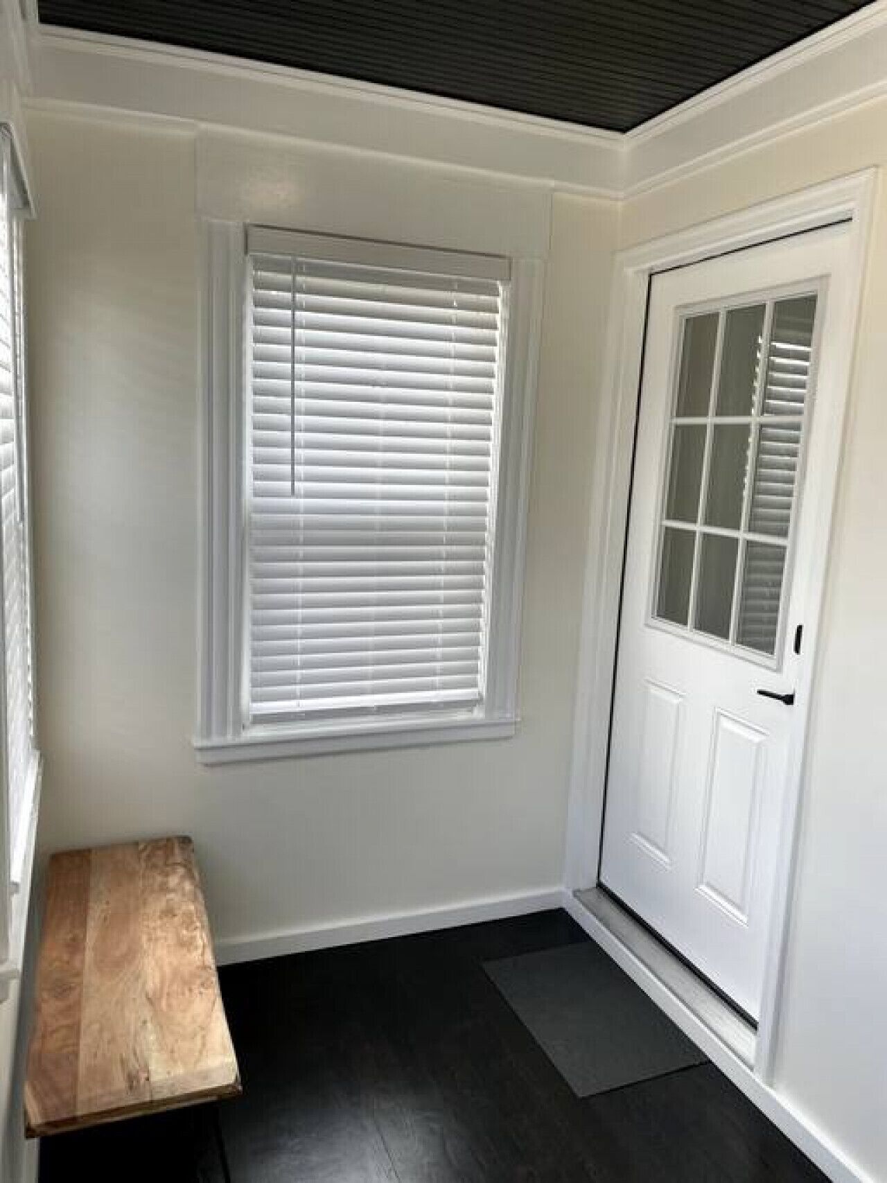 Een kamer met een witte deur, een raam met jaloezieën en een bankje.