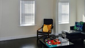 A living room with two windows, a black leather sofa, a wooden chair with a black cushion, and a coffee table with various items.