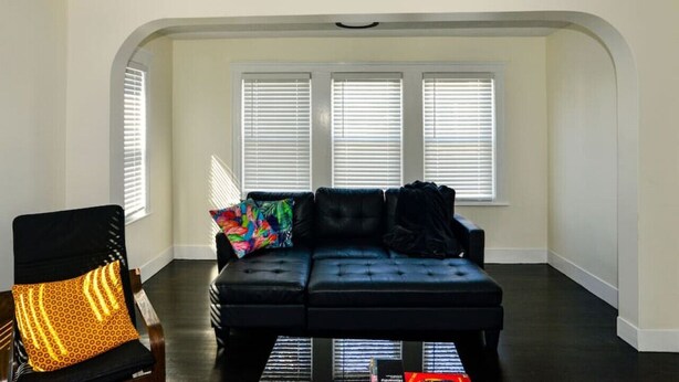 A living room with a black leather sofa, a patterned armchair, and a colorful throw pillow. There are three windows with blinds, and the room has a high ceiling.