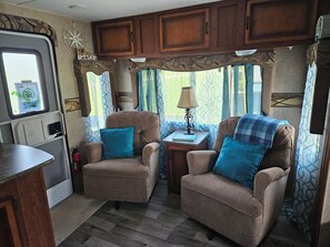 A compact RV interior with two upholstered chairs, a small table with a lamp, and a kitchenette area.