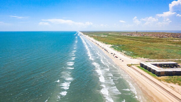 An aerial view of a beach with a long stretch of sand, a few buildings, and a clear sky. - Ocean View Condo (Corpus Christi)