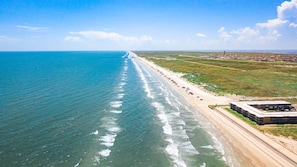 An aerial view of a beach with a long stretch of sand, a few buildings, and a clear sky. - Ocean View Condo (Corpus Christi)