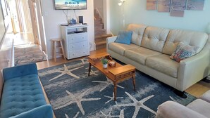 Un salon avec un canapé beige, un fauteuil bleu, une table basse et un téléviseur fixé au mur qui affiche « OCMD GETAWAYS ».