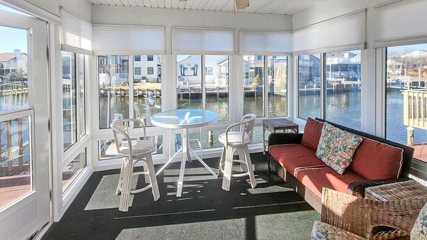 Une pièce lumineuse, baignée de soleil, avec une table et des chaises, un canapé, et une vue sur un plan d’eau et des bâtiments.