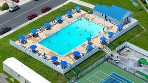 Vue aérienne d’un espace piscine avec des chaises longues et des parasols, entouré d’une clôture et d’un court de tennis.
