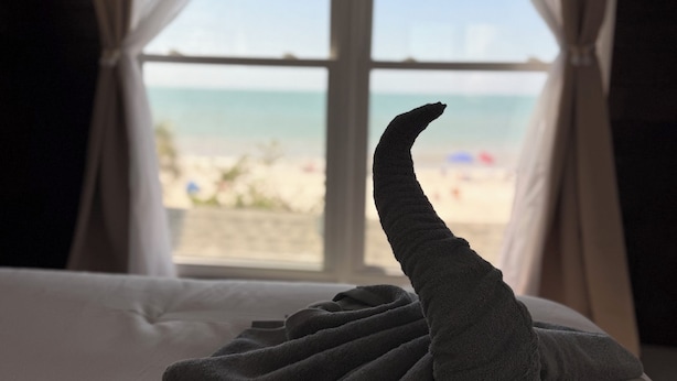 Ein Bett mit einem zu einem Schwan geformten Handtuch, ein Fenster mit Vorhängen und Blick auf einen Strand.