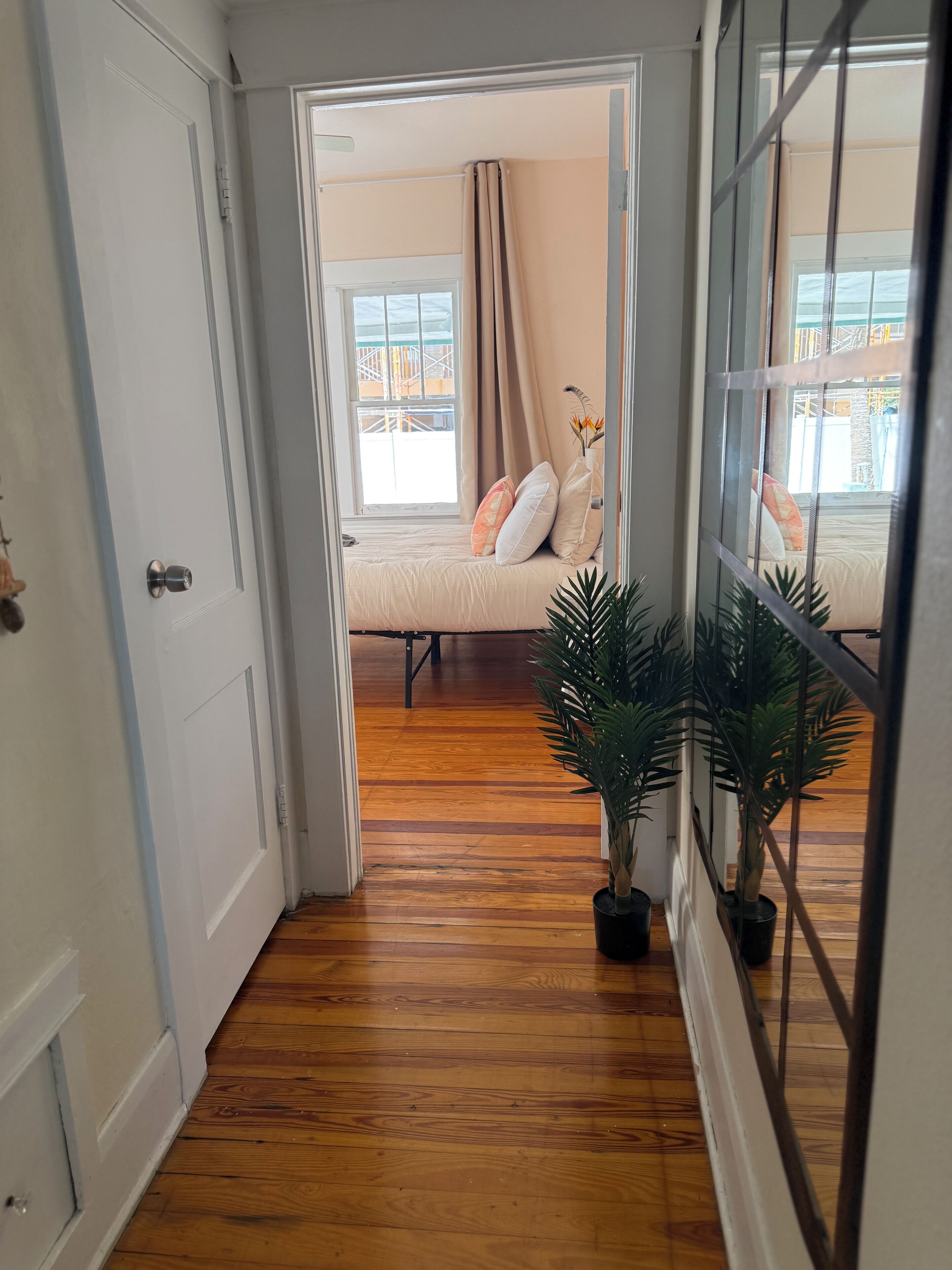 A hallway with wooden flooring leading to a bedroom with a bed, a window with curtains, and a potted plant.