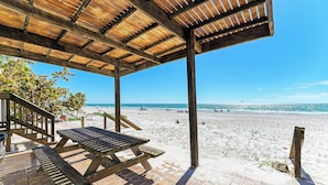 Ein hölzerner Strandpavillon mit Picknicktischen und einer gepflasterten Terrasse mit Blick auf einen Sandstrand und einen strahlend blauen Himmel.