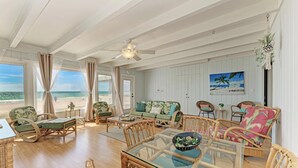 Ein geräumiges Wohnzimmer mit einem großen Glastisch, Korbmöbeln und Blick auf den Strand.