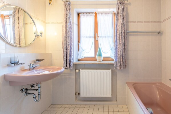 A bathroom with a pink sink, a bathtub, a window with curtains, and a radiator.