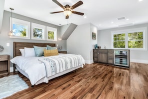 A bedroom with a wooden floor, a bed with a white and striped blanket, a ceiling fan, and a window with a view of trees.