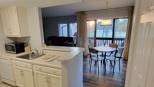 Eine moderne Küche mit weißen Schränken, einer eingebauten Mikrowelle und einer großen Kochinsel mit Spüle. Es gibt einen Essbereich mit Tisch und Stühlen sowie einen Blick ins Freie durch große Fenster.